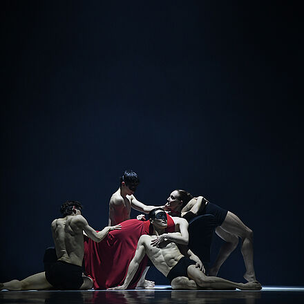 Eine Frau in Rot liegt auf dem Bett. Vor ihr knien Tänzer mit Masken.