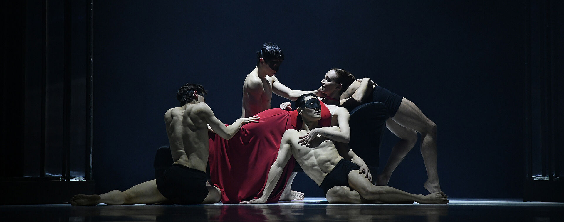 Eine Frau in Rot liegt auf dem Bett. Vor ihr knien Tänzer mit Masken.