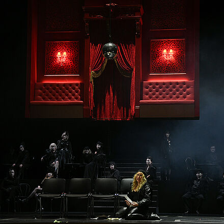 Im Vordergrund sitzt eine Frau auf dem Boden vor vier Stühlen. Im hinteren unteren Bereich des Bildes ist eine große, schwarz gekleidete Menschengruppe zu sehen. Im oberen Hintergrund schwebt ein Bühnenbild in der Luft.