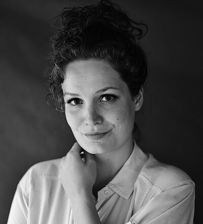 Schwarzweißes Portraitfoto von Annemarie Pfahler. Ihre linke Hand liegt auf ihrer rechten Schulter. Sie hat lockiges Haar, das sie zum Dutt hochgesteckt hat. Sie trägt eine helle Bluse.