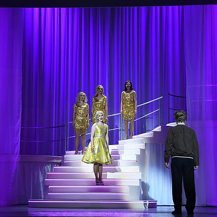 Eine junge Frau mit blonden Haaren und einem goldenen Kleid steht auf einer Showtreppe. Vor ihr steht ein junger blonder Mann. Er wendet sich vom Publikum ab und blickt auf die junge Frau. Hinter ihr stehen drei weitere Frauen in goldenen Glitzerkleidern.
