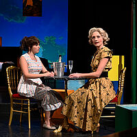 Lotte Körner sitzt gemeinsam mit Irene Gerlach am Tisch. Darauf steht ein Sektkühler mit einer Flasche darin sowie ein gefülltes Sektglas. Luise schaut betrübt.