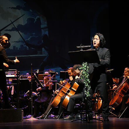 Im Licht steht eine Person, im Hintergrund befindet sich das Orchester.  