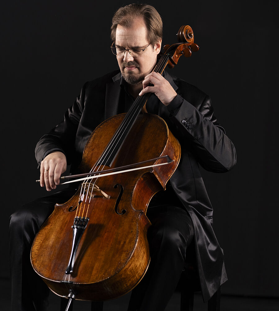 Foto von Cellist Wolfgang Emanuek Schmidt. Er hat braune kurze  Haare und einen Bart um den Mund. Er trägt einen schwarzen Anzug mit schwarzem Hemd und eine Brille.  Er sitzt und spielt Cello. Dabei blickt er nach unten auf die Cellosaiten.