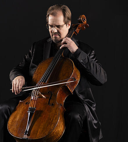 Foto von Cellist Wolfgang Emanuek Schmidt. Er hat braune kurze  Haare und einen Bart um den Mund. Er trägt einen schwarzen Anzug mit schwarzem Hemd und eine Brille.  Er sitzt und spielt Cello. Dabei blickt er nach unten auf die Cellosaiten.