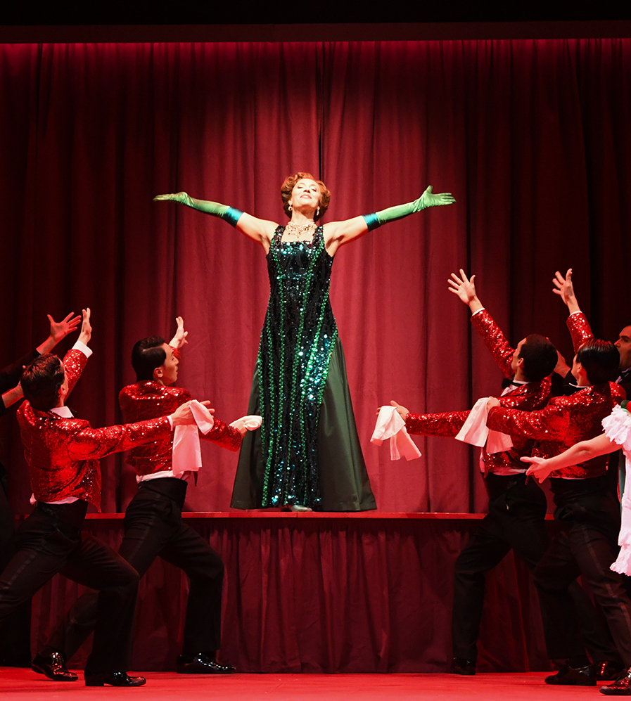 Die Bühne ist rot. Im Vordergrund stehen Tänzerinnen und Tänzer, während Gäste an Tischen sitzen. Auf der Bühne befindet sich eine Frau in einem grünen Glitzerkleid.