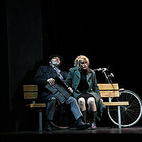Bess McNeill und ein Mann sitzen auf einer Parkbank. Dahinter lehnt ein Fahrrad. Sie schaut starr nach vorn. Er hat seine Augen geschlossen und hält eine Aktentasche auf dem Schoß. 