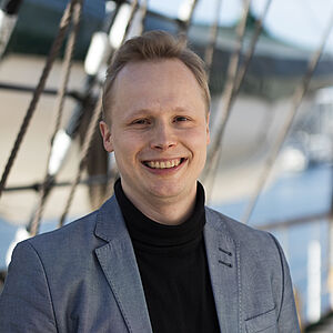 Portraitfoto von Franz Berlin. Er steht vor Tauen und lächelt mit geöffnetem Mund. Er trägt einen grauen Blazer und einen schwarzen Rollkragenpullover. Sein Haar ist blond und nach oben gegelt.