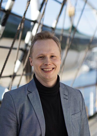 Portraitfoto von Franz Berlin. Er steht vor Tauen und lächelt mit geöffnetem Mund. Er trägt einen grauen Blazer und einen schwarzen Rollkragenpullover. Sein Haar ist blond und nach oben gestylt.