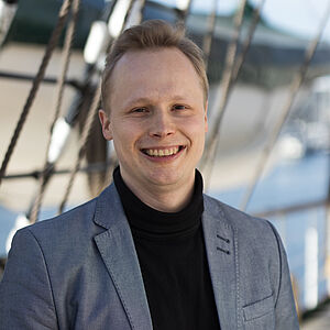 Portraitfoto von Franz Berlin. Er steht vor Tauen und lächelt mit geöffnetem Mund. Er trägt einen grauen Blazer und einen schwarzen Rollkragenpullover. Sein Haar ist blond und nach oben gestylt.
