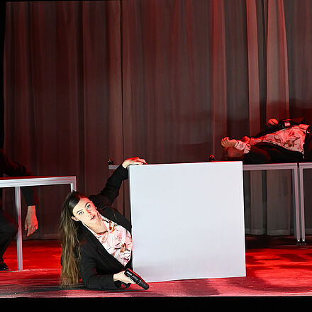 Links im Bild sitzt ein Mann an einem Tisch. Er liegt mit dem Kopf auf der Tischplatte. In der Mitte des Bildes liegt ein Tisch auf dem Boden. Hinter der Tischplatte liegt eine Frau, die einen Akkuschrauber in der Hand hält und nach vorne schaut. Rechts im Bild stehen zwei Tische, auf denen ein Mann liegt.