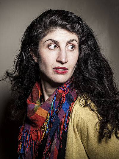 Portraitfoto von Carolin Löffler. Sie hat langes gewelltes dunkles Haar. Sie trägt einen senffarbenen Pullover und einen buntgemusterten Schal.