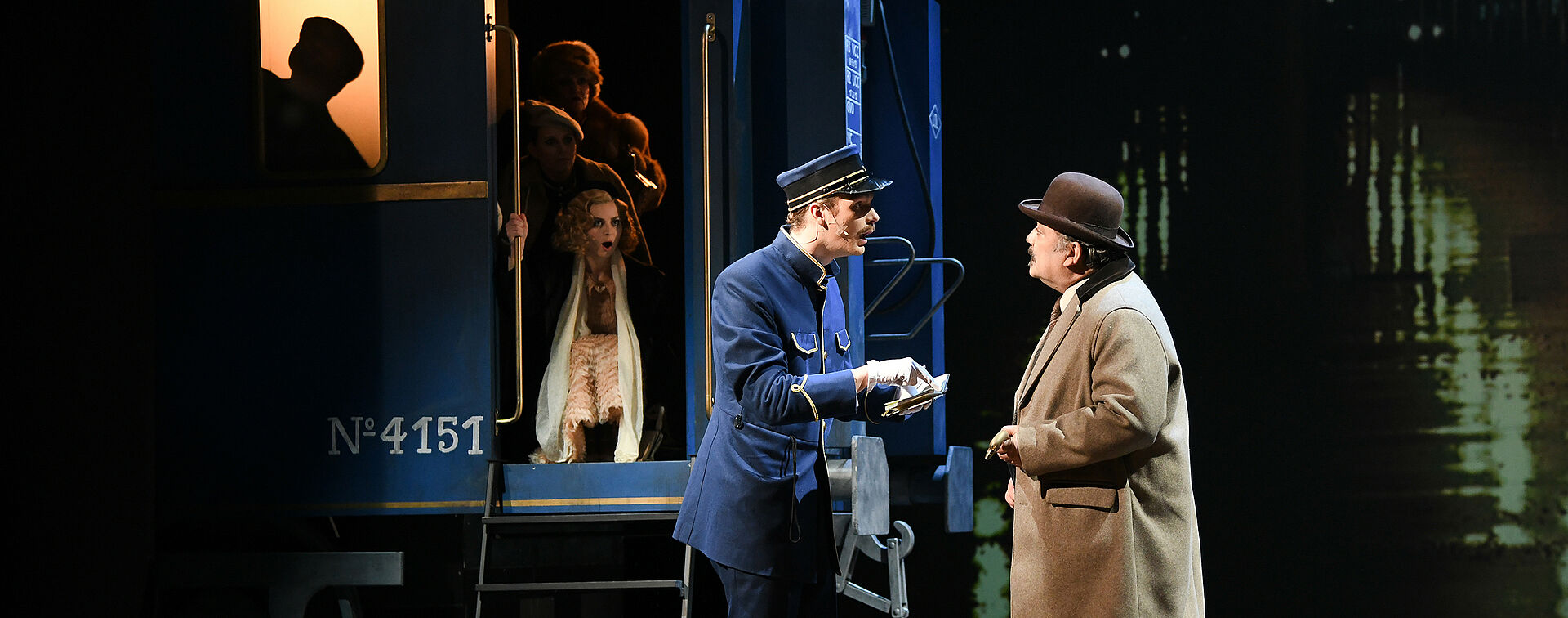 Michel in Schaffner-Uniform und Hercule Poirot mit Koffern und Hut (Melone) im Gespräch. Michel deutet mit geöffnetem Mund auf das Büchlein in seinen Händen. Andere Passagiere schauen aus einem blauen Zug heraus auf die Szenerie.