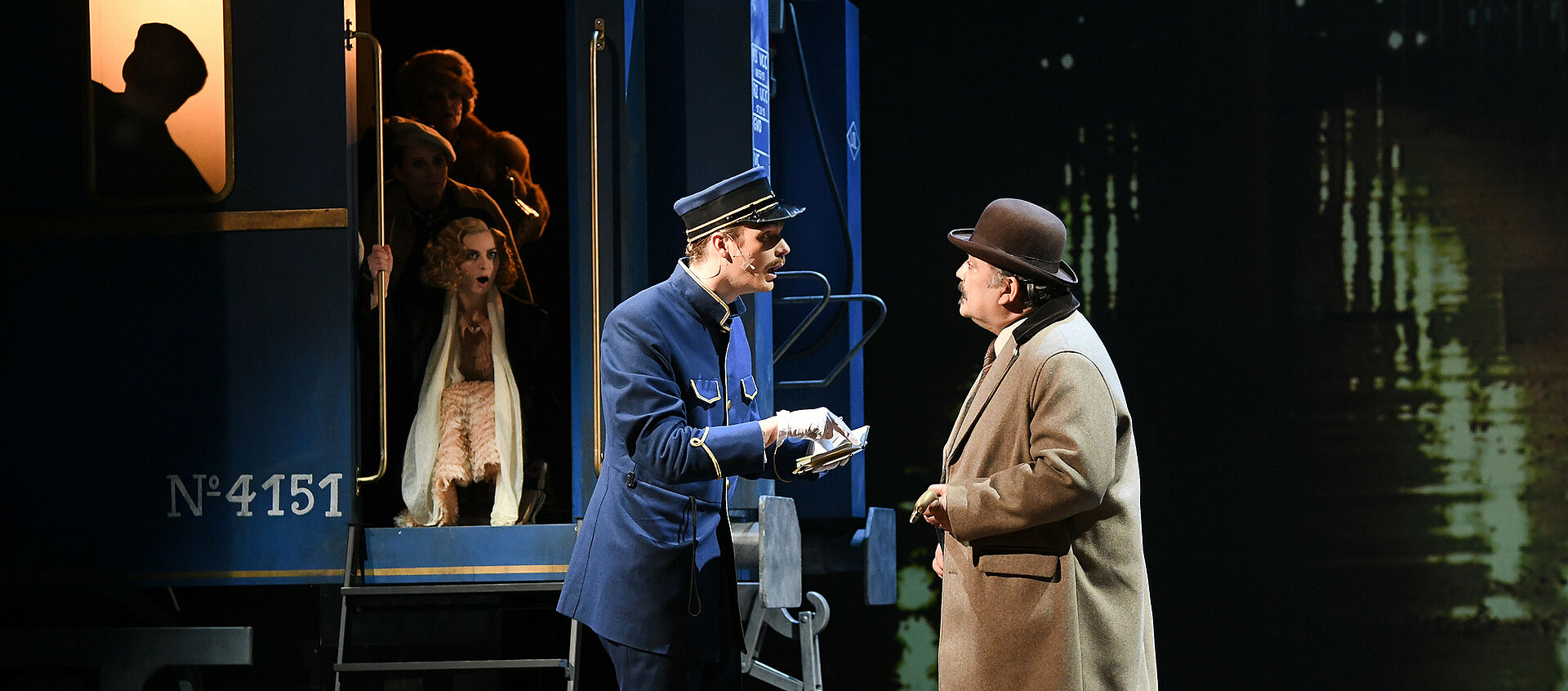 Michel in Schaffner-Uniform und Hercule Poirot mit Koffern und Hut (Melone) im Gespräch. Michel deutet mit geöffnetem Mund auf das Büchlein in seinen Händen. Andere Passagiere schauen aus einem blauen Zug heraus auf die Szenerie.