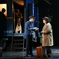 Michel in Schaffner-Uniform und Hercule Poirot mit Koffern und Hut (Melone) im Gespräch. Michel deutet mit geöffnetem Mund auf das Büchlein in seinen Händen. Andere Passagiere schauen aus einem blauen Zug heraus auf die Szenerie.