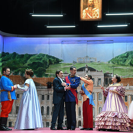 Viele Personen in Anzügen und opulenten Kleidern stehen auf der Bühne. Einige halten sich gegenseitig fest oder richten ihre Kleidung.