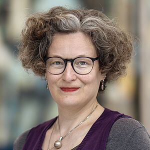 Portraitfoto von Bianca Sue Henne. Sie hat lockiges volles Haar, welches sie kinnlang trägt. Es ist braun-gräulich. Sie trägt eine Brille und roten Lippenstift sowie Schmuck. Sie ist in einen grauen Pullover unter einem lilafarbenen Pullunder gekleidet. Der Hintergrund ist verschwommen.