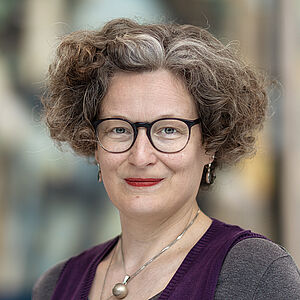 Portraitfoto von Bianca Sue Henne. Sie hat lockiges volles Haar, welches sie kinnlang trägt. Es ist braun-gräulich. Sie trägt eine Brille und roten Lippenstift sowie Schmuck. Sie ist in einen grauen Pullover unter einem lilafarbenen Pullunder gekleidet. Der Hintergrund ist verschwommen.