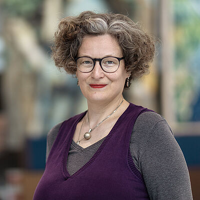 Portraitfoto von Bianca Sue Henne. Sie hat lockiges volles Haar, welches sie kinnlang trägt. Es ist braun-gräulich. Sie trägt eine Brille und roten Lippenstift sowie Schmuck. Sie ist in einen grauen Pullover unter einem lilafarbenen Pullunder gekleidet. Der Hintergrund ist verschwommen.