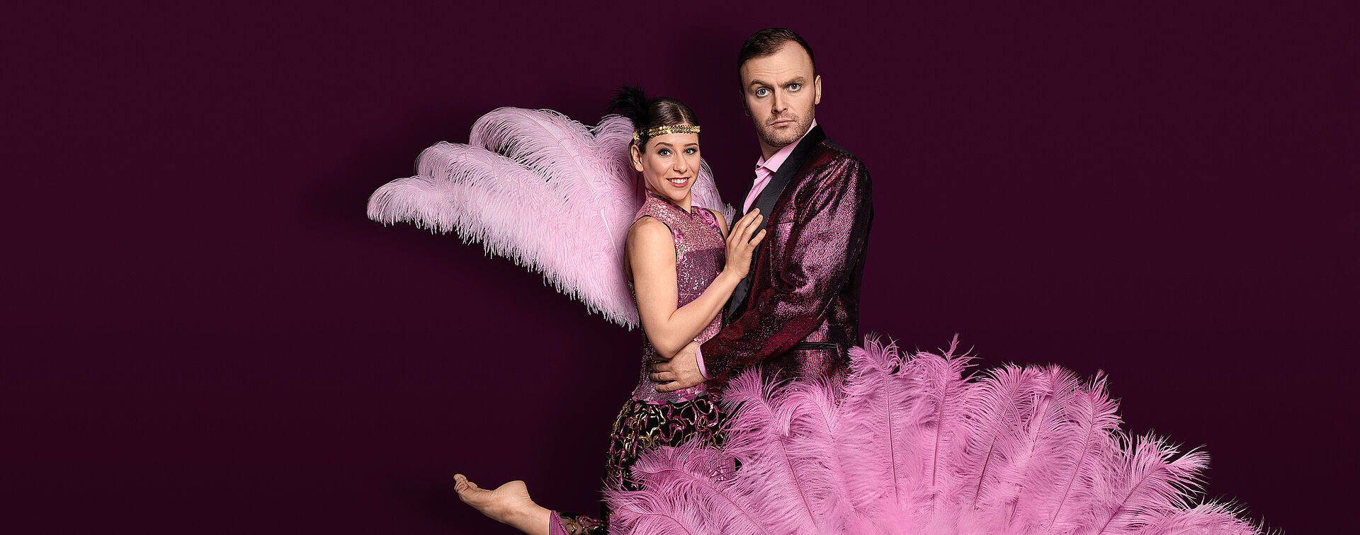 Eine Frau (Melissa Panetta) und ein Mann (Andrew Irwin) stehen in einer Paar-Umarmung zu einander. Vor ihnen ist ein rosa Federfächer und hinter ihrem Rücken ist ebenfalls ein kleiner rosa Federfächer. Der Hintergrund ist weinrot.  