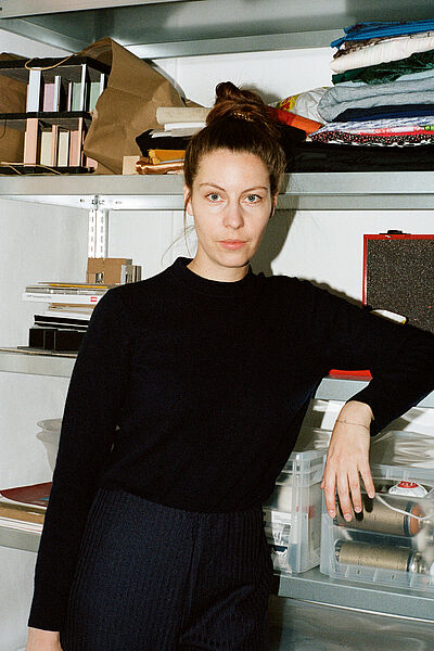 Portraitfoto von Kathrin Kemp. Sie seht umgeben von gefüllten weißen Regalflächen und lehnt auf einer gefüllten transparenten Aufbewahrungsbox aus Plastik. Ihr Haar ist auf dem Kopf zu einem Knoten gebunden. Sie trägt einen schwarzen Pullover, der in einer dunkelblauen gestreiften Hose steckt.