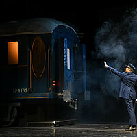 Schaffner Michel trägt blaue Uniform. Er ist zum Zug gedreht und hat seine linke Hand gehoben, im Mund eine Trillerpfeife. Vor ihm ein dampfender Zugwaggon.