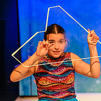 Die Schauspielende malt mit ihrem Finger Wellen auf die von ihrem Atem angehauchte Glasscherbe, welche sie vor ihrem Kopf emporhält. Sie trägt ein buntes Top. 