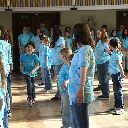 Der Kinderchor steht mit den Körpern nach links gedreht in mehreren Reihen und mit ausgestreckten Armen. Zwischen den Reihen läuft ein Kind. Alle tragen ein blaues Oberteil.