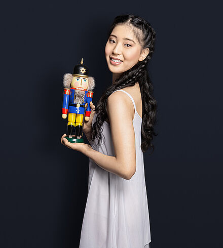 Eine Frau mit langen schwarzen Haaren, die offen geflochten sind, hält inn ihren Händen einen blauen Nussknacker. Sie trägt ein weißes Kleid und lächelt. Der Hintergrund des Bildes ist dunkelblau.