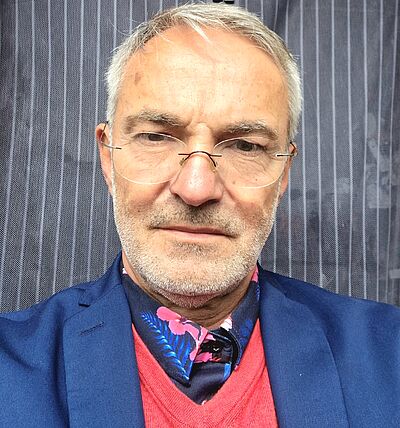 Portraitfoto von Dorin Gal. Er trägt ein blaues Jackett, darunter einen roten Pollover und ein geblümtes Hemd. Sein Haar ist grau. Er trägt eine randlose Brille. Dei Hintergrund ist längs gestreift.