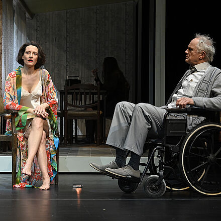 Eine Frau im Morgenmantel sitzt auf einem Stuhl, auf dem Schoß ein Buch. Neben ihr sitzt ein Mann im Rollstuhl. Im Hintergrund erkennt man eine Frau, die am Frühstückstisch sitzt.