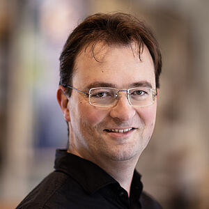 Portraitfoto von Jorrit van den Ham. Er steht seitlich. Er trägt braunes Haar, eine randlose eckige Brille und ein schwarzes Hemd.