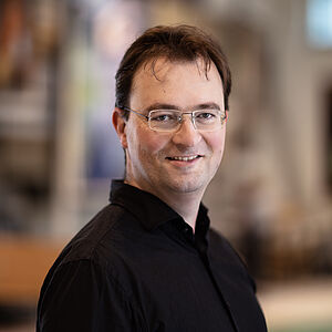 Portraitfoto von Jorrit van den Ham. Er steht seitlich. Er trägt braunes Haar, eine randlose eckige Brille und ein schwarzes Hemd.