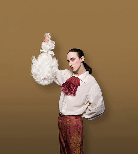 Mann in barockem Kostüm hält eine Perücke empor
