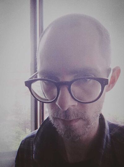 Portraitfoto von David Loscher. Er steht vor einem Fenster. Er trägt sein Haar kurzgeschoren, einen Dreitagebart und eine Brille. Sein Hemd ist kariert. Er schaut über den Rand der Brille. Das Bild ist von einem weißen Nebel überzogen.