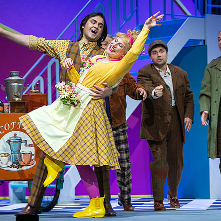 Ein Mann mit karierter Weste tanzt mit der jungen Frau in gelben Kleidern (Eliza Doolittle). Im Hintergrund steht ein Gruppe Männer in einfachen Klamotten, sie singen.