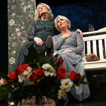 Zwei Frauen sitzen Arm in Arm auf der Bank. Im Vordergrund sitz Pflanzen zu sehen 