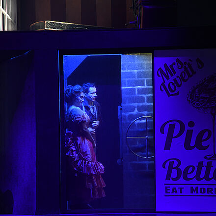 Im Mittelpunkt steht eine Frau und ein Mann an einer Wand, sie schauen in eine Richtung. Auf dem Schild davor kann man «Pie makes it all better. Eat more Pie»