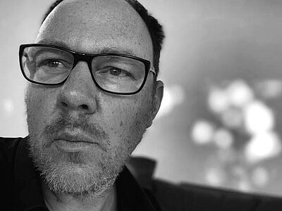 Schwarzweißes Portraitfoto von Frank Albert. Er schaut von oben skeptisch in die Kamera. Er trägt eine Brille mit dunklem Rahmen, einen Dreitagebart sowie ein schwarzes Hemd. Der Hintergrund ist verschwommen.