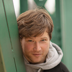 Portraitfoto von Marc Vinzing. Er lehnt sich auf den Streben einer grünen Stahlbrücke. Er steht geduckt und schaut hinter den Streben hervor. Er hat vollen helles dunkelblondes Haar, trägt einen Dreitagebart, einen hellgrauen Hoodie und darüber einen schwarzen Mantel.
