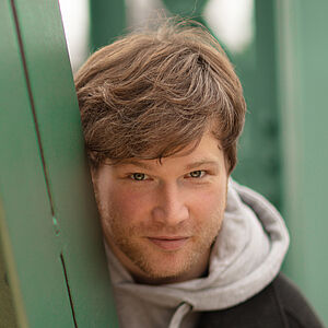 Portraitfoto von Marc Vinzing. Er lehnt sich auf den Streben einer grünen Stahlbrücke. Er steht geduckt und schaut hinter den Streben hervor. Er hat vollen helles dunkelblondes Haar, trägt einen Dreitagebart, einen hellgrauen Hoodie und darüber einen schwarzen Mantel.