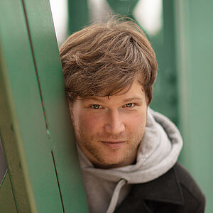 Portraitfoto von Marc Vinzing. Er lehnt sich auf den Streben einer grünen Stahlbrücke. Er steht geduckt und schaut hinter den Streben hervor. Er hat vollen helles dunkelblondes Haar, trägt einen Dreitagebart, einen hellgrauen Hoodie und darüber einen schwarzen Mantel.