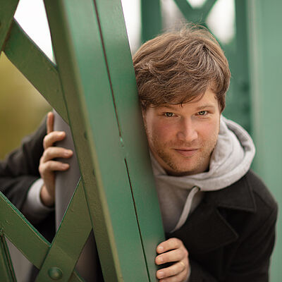 Portraitfoto von Marc Vinzing. Er lehnt sich auf den Streben einer grünen Stahlbrücke. Er steht geduckt und schaut hinter den Streben hervor. Er hat vollen helles dunkelblondes Haar, trägt einen Dreitagebart, einen hellgrauen Hoodie und darüber einen schwarzen Mantel.