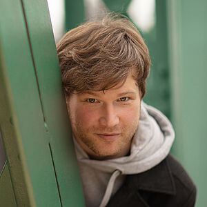 Portraitfoto von Marc Vinzing. Er lehnt sich auf den Streben einer grünen Stahlbrücke. Er steht geduckt und schaut hinter den Streben hervor. Er hat vollen helles dunkelblondes Haar, trägt einen Dreitagebart, einen hellgrauen Hoodie und darüber einen schwarzen Mantel.