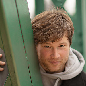 Portraitfoto von Marc Vinzing. Er lehnt sich auf den Streben einer grünen Stahlbrücke. Er steht geduckt und schaut hinter den Streben hervor. Er hat vollen helles dunkelblondes Haar, trägt einen Dreitagebart, einen hellgrauen Hoodie und darüber einen schwarzen Mantel.