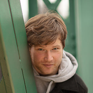 Portraitfoto von Marc Vinzing. Er lehnt sich auf den Streben einer grünen Stahlbrücke. Er steht geduckt und schaut hinter den Streben hervor. Er hat vollen helles dunkelblondes Haar, trägt einen Dreitagebart, einen hellgrauen Hoodie und darüber einen schwarzen Mantel.