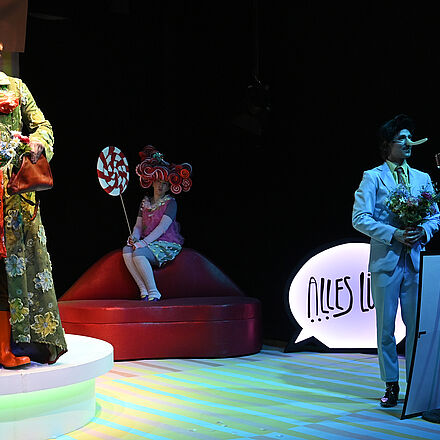 Links im Bild steht eine Person auf einem Podest. In der Mitte des Bildes sitzt eine Frau auf einem lippenförmigen Sofa. Rechts im Bild steht ein Mann mit einer langen Nase vor einem Mikrofon.