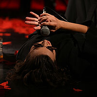 Eine Sängerin mit Sonnenbrille liegt am Boden. Sie singt konzentriert in ein Mikrofon. Neben ihr steht eine Flasche Whisky.