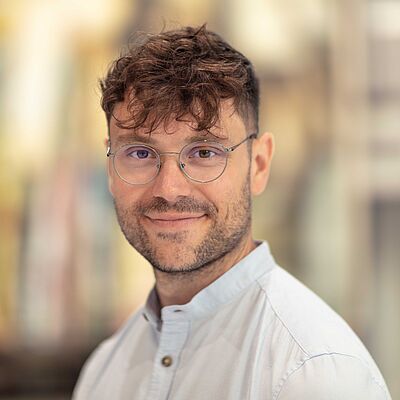 Das Porträt zeigt Edward Münch, einen Mann mit braunem Haar. Er trägt ein weißes Hemd und eine Brille und steht lächelnd vor einem sanft unscharfen Hintergrund.