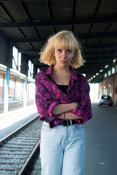 Portraitfoto von Fenja Abel. Sie läuft neben einem Gleisbett in einem Bahnhof entlang. Ihre Arme sind verschränkt. Sie trägt eine lilafarbene gemusterte Bluse zu einer hellen Jenas. Sie hat blondes schulterlanges Haar mit Pony.